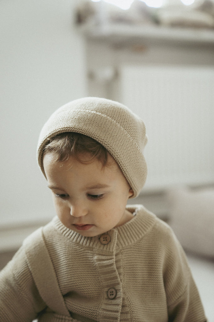 Goldiges Kind trägt eine Mütze aus feiner Baumwolle in beige und einen Strickcardigen mit einer Latzhose