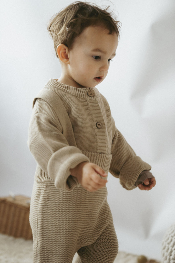 Kleinkind mit einem beigefarbenen Outfit bestehend aus einem Cardigan und einer Latzhose aus feiner Bio-Baumwolle 
