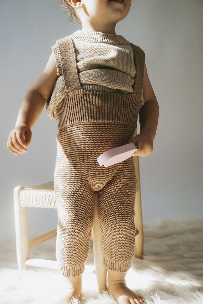 Niedliches Kind trägt eine stylische braune Latzhose aus feiner Baumwolle mit einem beigen Pullunder. Es hat ein rosa Eis in der Hand