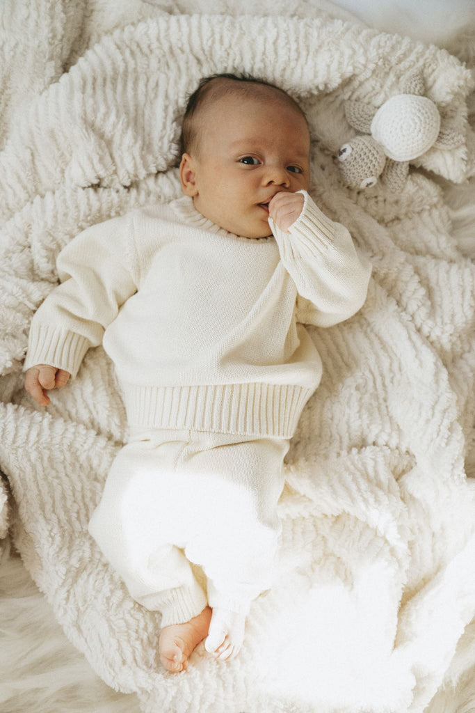 Süßes Baby mit Daumen im Mund trägt einen Strickpullover aus feiner Baumwolle in Creme