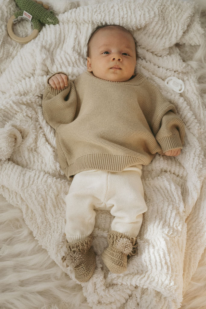Niedliches Baby liegt auf einer Decke und trägt Klamotten aus Biobaumwolle in Beige und Creme