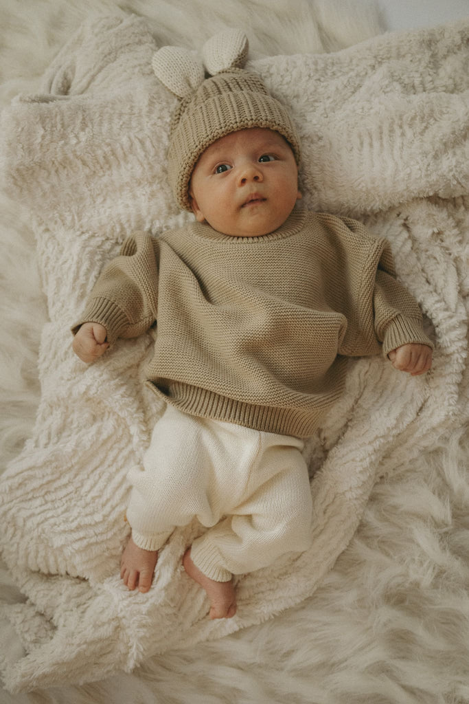 Süßes Baby mit goldiger Wollmütze mit Hasenohren und einem beigefarbenen Pullover aus Baumwolle trägt eine Creme Strickhose