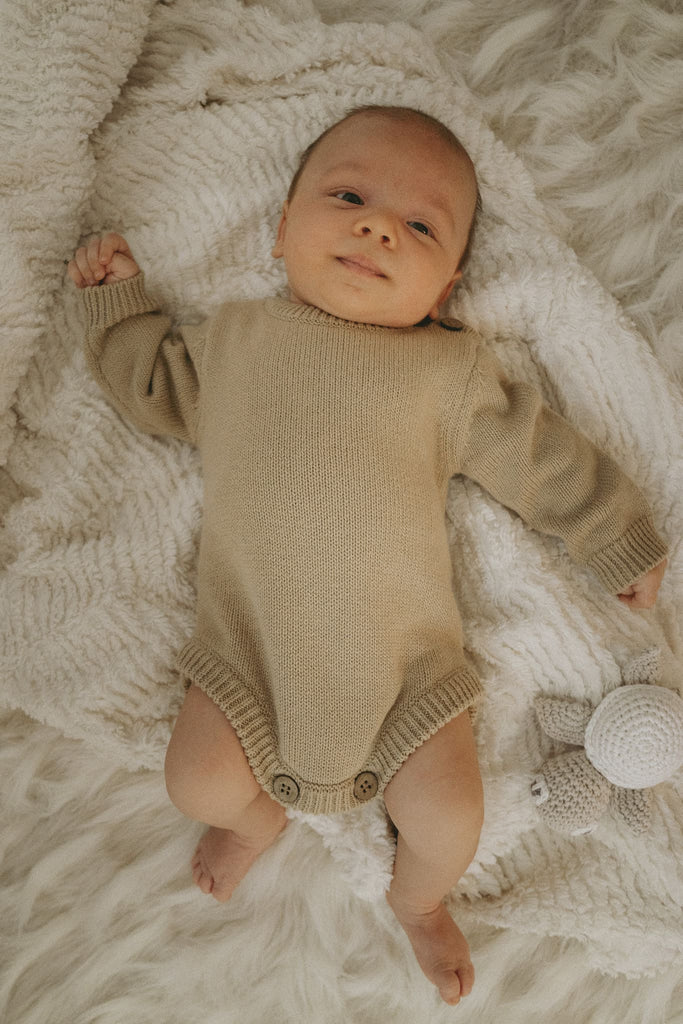 Baby mit einen beigen Body aus Bio-Baumwolle liegt grinsend auf einem weichen Teppich